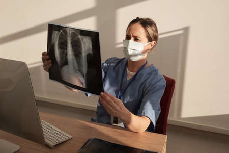 Young female radiologist in protective mask looking at x-ray image of patient while sitting by desk in front of computerの写真素材