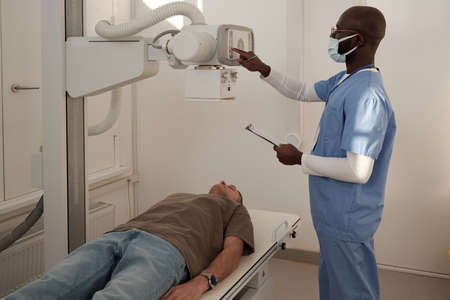 Young clinician in uniform and protective mask turning on x-ray machine before examining mature male...