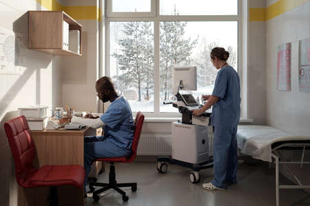 Two young intercultural clinicians preparing for ultrasound examination while man making notes and...