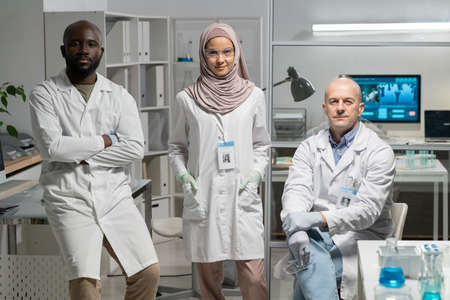 Group of three interracial microbiologists in lab coats sitting by workplace in clinical or...