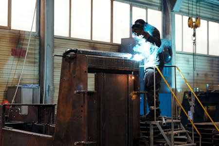 Contemporary worker or welder in protective mask and uniform using arc welding while repairing huge...