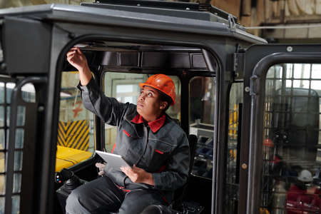 African American female worker of industrial plant with tablet driving construction machine while...