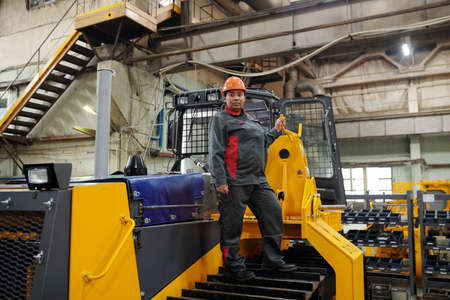 Young confident female worker standing on caterpillar of construction machine and holding by handle...