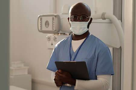Contemporary male clinician in protective mask with medical document standing against roentgen...