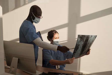 Young African American assistant in uniform and protective mask pointing at x-ray image held by female radiologistの写真素材