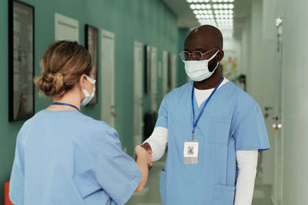 Young African American surgeon in uniform and protective mask shaking hand of his female assistant while both standing in hospital corridorの写真素材