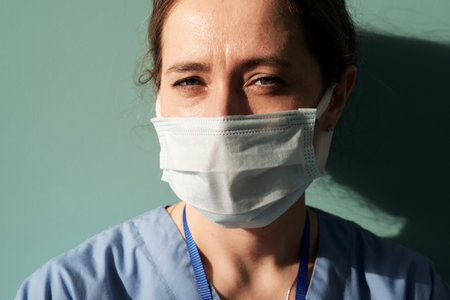 Face of young female general practitioner or assistant of surgeon in protective mask looking at camera while standing by wallの写真素材