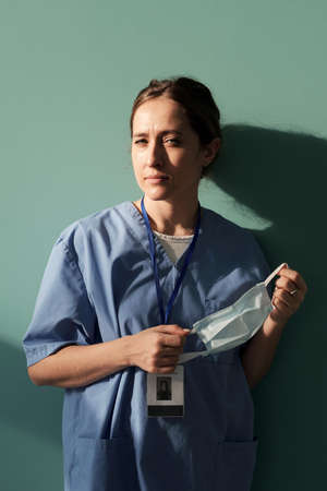 Young female assistant or physician in blue uniform holding protective mask in hands while standing by wall of hospital wardの写真素材