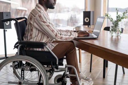 Young African American freelancer with disability typing in front of laptop while sitting by wooden...