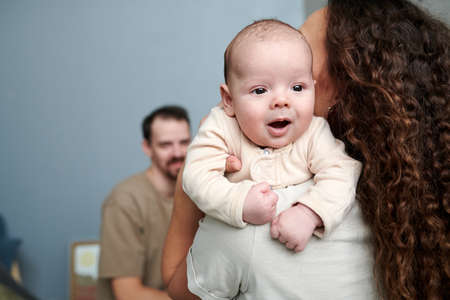 Happy adorable baby boy in white romper suit on hands of his mother standing in front of her husband at homeの写真素材