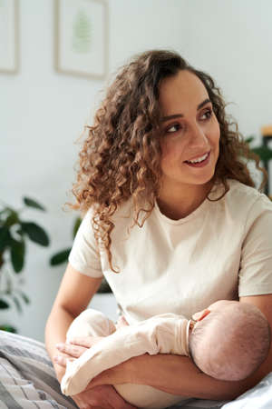 Happy young female with dark long wavy hair holding sleeping baby son on hands while communicating with someoneの写真素材