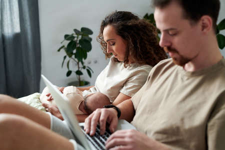 Young serene woman with newborn baby son on hands sitting on bed by her husband typing on laptop keyboard while networkingの写真素材