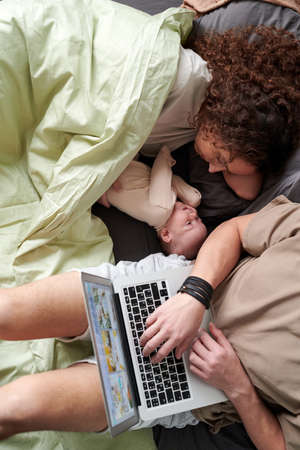 Above angle of young businessman typing on laptop keyboard while sitting on bed next to his wife lulling their adorable baby sonの写真素材
