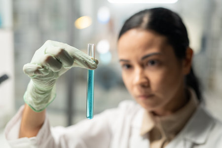 Gloved hand of young female scientist in lab coat holding flask with blue liquid while carrying out...