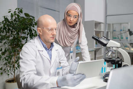 Two young contemporary virologists discussing online data in laboratory whilemature male scientist pointing at tablet screenの写真素材