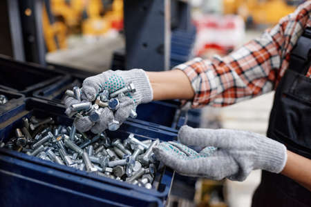 Gloved hands of female engineer in workwear taking pile of bolts out of plastic box while standing in front of display with building materialsの写真素材