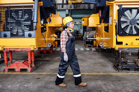 Young female technician in overalls, harhat and gloves moving along large industrial machines standing in rows in warehouseの写真素材