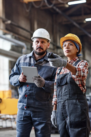 Engineers of large contemporary industrial plant looking at huge machine while female regulating its...