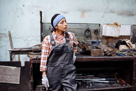 Young contemporary repairperson in coveralls standing by workbench with open toolkit and variety of worktools on shelvesの写真素材