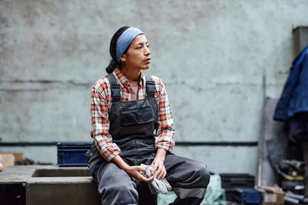 Young tired female engineer in coveralls and headband taking short rest after hard work in large warehouse of modern factory or plantの写真素材