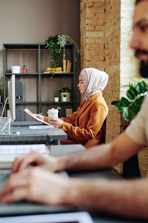 Young Muslim female accountant in hijab and casual clothes looking through financial papers while sitting by workplaceの写真素材