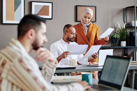 Two young Muslim coworkers looking through financial documents by workplace in front of male broker sitting in front of laptopの写真素材