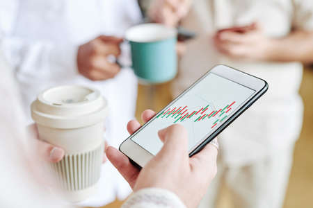 Young contemporary businessman with cup of coffee analyzing chart in smartphone while standing in...