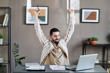 Young successful businessman expressing joy in front of laptop whie sitting with his arms raised by desk in office or bureauの写真素材