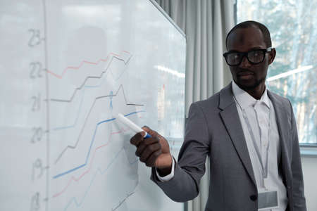 Young serious African American teacher pointing at income graph on whiteboard during presentation of company incomeの写真素材