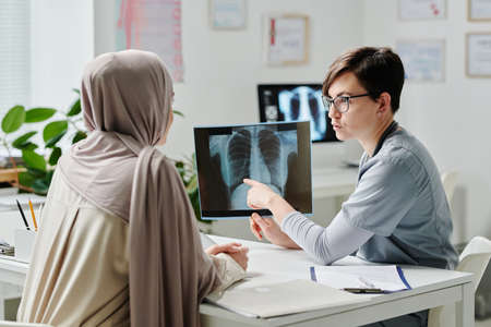 Confident female radiologist showing result of medical examination to patient in hijab while sitting...