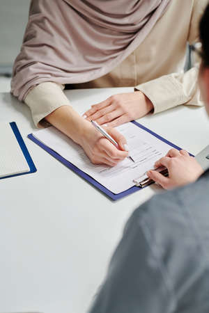 Hand of young Muslim female patient with pen pointing at medical form on clipboard while filling in...