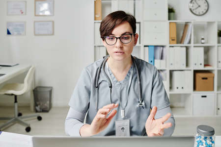 Confident doctor in medical scrubs communicating to online patients while sitting by desk in office...