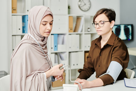 Young Muslim female physician in hijab checking blood pressure of patient while sitting by workplace...