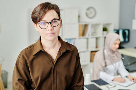 Young brunette female patient in eyeglasses and brown casual shirt looking at camera while standing...
