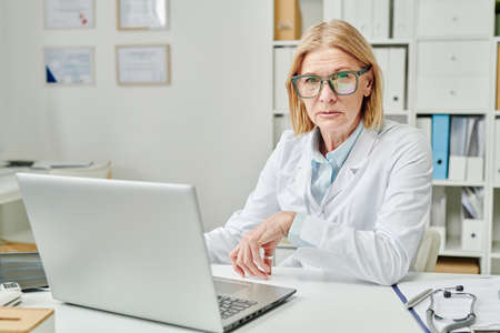 Confident mature experienced doctor in whitecoat sitting by workplace in front of laptop and looking at camera during online consultationの写真素材