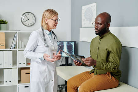Young African American male patient looking at x-ray of his lungs while sitting in hospital and listening to explanation of mature doctorの写真素材