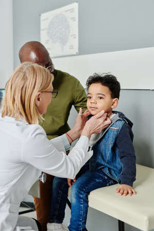 Experienced mature female endocrinologist in whitecoat checking thyroid of cute little patient visiting clinics with his fatherの写真素材