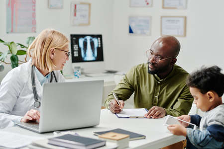 Experienced doctor with laptop consulting African American male patient filling in medical form...