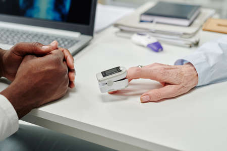 Hand of mature female patient with pulse oximeter on fingertip sitting in front of doctor during...