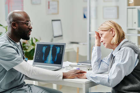 Young doctor comforting mature female patient with dangerous disease while sitting by workplace in...
