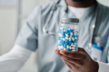 Hand of young African American male physician holding plastic jar of pills while showing it to...