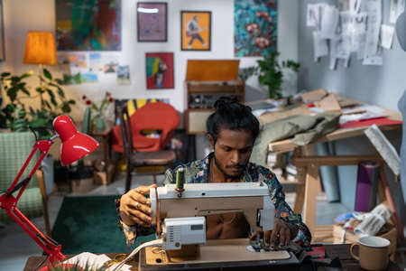 High angle shot of creative young fashion designer of Indian ethnicity sitting at table using sewing machine to make some new clothesの写真素材