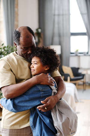 Senior black man embracing his cute grandson with backpack in living room of loft apartment while seeing him off for schoolの写真素材