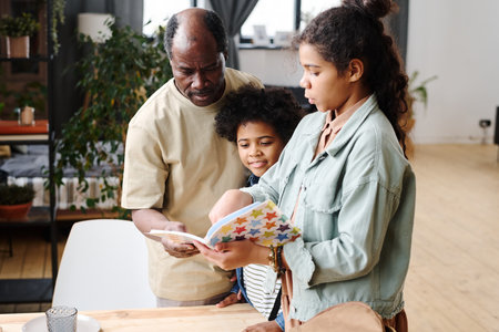 Contemporary senior black man helping his granddaughter with home assignment written in copybook while looking at her notesの写真素材