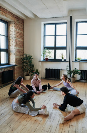 Group of active teens sitting on wooden floor of spacious dance hall or studio and practicing stretching and balance exercisesの写真素材