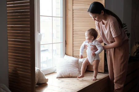 Modern young mother spending time at home with her lovely daughter teaching her to stand and walk