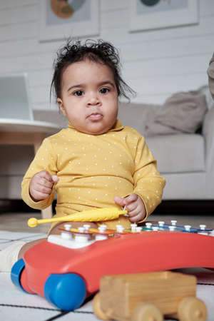Vertical portrait of lovely African American baby wearing yellow clothes sitting on floor playing...