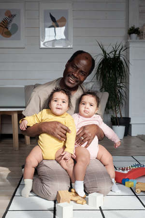 Portrait of cheerful mature African American man sitting on floor in living room at home hugging twin babies smiling at cameraの写真素材