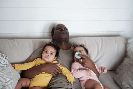 High angle view of exhausted mature African American dad falling asleep while taking care of his...
