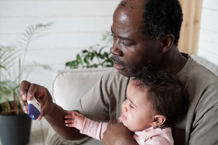 Medium close-up of mature Black man checking body temperature of his baby using electronic...
